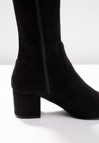 Černá semišová kotníková bota s kulatou špičkou, masivním podpatkem a bočním zipem. Texturovaný povrch a minimalistické designové prvky.