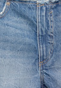 Nära bild av blå denimbyxor som visar framfickan, blixtlåssömmen, kopparniten och detaljerna med orange sömnad.