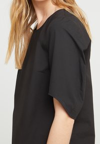 Schwarzes Kurzarmshirt mit Rundhalsausschnitt und seitlicher Tasche. Der Stoff scheint glatt und leicht zu sein, mit einem lässigen Schnitt.