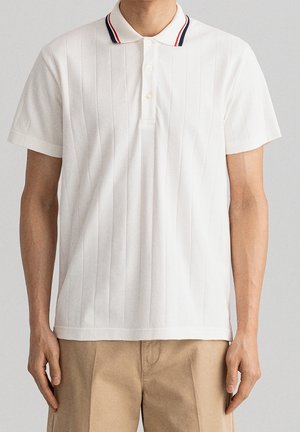 Mann trägt weißes Poloshirt mit kurzen Ärmeln und rot-navy gestreiftem Kragen, kombiniert mit beigen Hosen, steht vor schlichtem Hintergrund.
