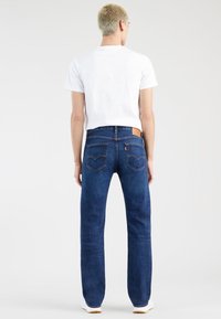 Personne aux cheveux blonds courts, de dos, portant un t-shirt blanc, un jean bleu droit et des baskets blanches sur un fond uni.