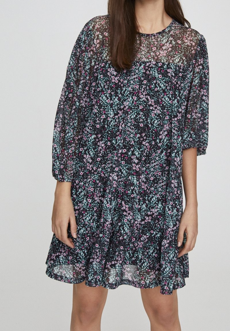 Černé květinové šaty vyrobené z průhledné látky, s kulatým výstřihem, dlouhými nafouklými rukávy a zvonovým spodním lemem s barevnými květinovými vzory.