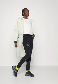 Kvinne iført en lett lang frakk, hvit skjorte, svarte leggings med "Dynafit"-logo, gule sokker og svarte joggesko, stående med den ene hånden i lomma.