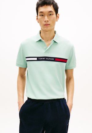 Giovane uomo con polo verde chiaro Tommy Hilfiger con striscia logo sul petto e pantaloni scuri, in piedi con le mani in tasca.