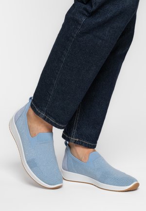 Pies con zapatillas sin cordones en azul claro con suelas blancas y detalles de brillo, combinadas con vaqueros de mezclilla azul oscuro.