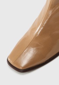 Beige kožené kotníkové boty s lesklým povrchem, špičatou špičkou a minimem švů, které představují elegantní a moderní design.