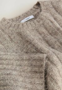 Pull en tricot beige avec une texture côtelée, présentant un col rond et une étiquette en tissu indiquant la marque "NEXT."