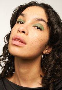 Portrait en gros plan d'une personne avec des cheveux noirs bouclés, portant un fard à paupières vert, du rouge à lèvres clair et de petites boucles d'oreilles en argent.