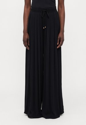 Persoon draagt zwarte wijde broek met aantrekkoord in de taille en een aansluitende zwarte top tegen een effen achtergrond.