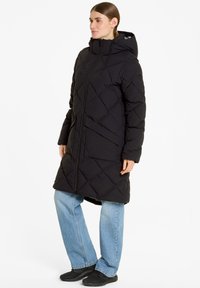 Piumino nero con zip frontale, cappuccio e pattern trapuntato. Presenta tasche laterali e un taglio fino al ginocchio, abbinato a jeans azzurri chiari.