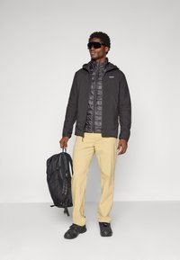 Veste isolante noire sur une couche thermique sombre, pantalon beige et chaussures noires. La personne porte un sac à dos noir avec des bretelles réglables.