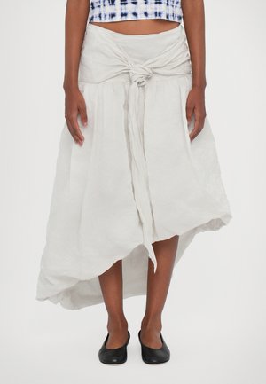 White linen skirt with a knotted waist detail, asymmetrical hem, and soft, textured finish. Paired with simple black slip-on shoes.