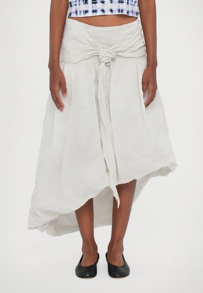 Witte linnen rok met een geknoopte taille, asymmetrische zoom en een zachte, textuurachtige afwerking. Gecombineerd met eenvoudige zwarte instappers.