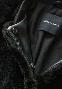 Svart imitation pälskrage med en slät läderknapp. Inuti har den ett matt svart tygfoder och en kläklapp sydd på sidan.