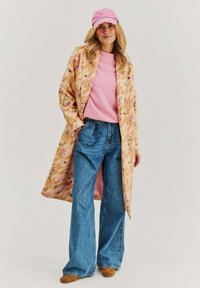 Světlý hnědý dlouhý kabát s květinovým vzorem, růžový svetr, modré široké džíny a růžová kšiltovka. Výrazné texturované látky a barvy.