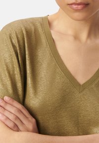 Top v-neck in verde oliva metallizzato con texture fine, maniche corte e bordi a costine. Elegante caduta sulle braccia incrociate, che mette in risalto il décolleté.