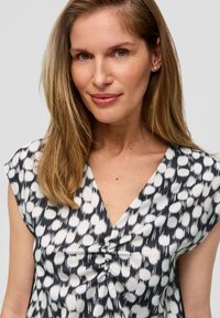 V-hals blouse met een zwart-wit abstract patroon, korte mouwen en een geplooid detail aan de voorkant. Gladde stoffen textuur.