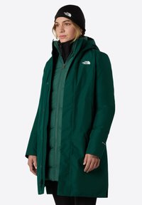 Grøn isoleret parka med hætte, der har lynlås foran, trykknapper og logo-detaljer. Lagdelt design med en glat tekstur.