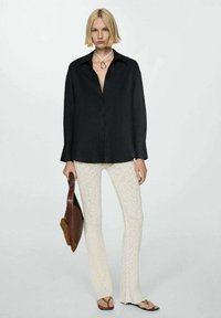 Camasa neagră cu guler, asortată cu pantaloni crem cu dungi și o geantă de umăr maro. Modelul poartă șlapi și un colier simplu.