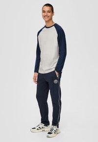 Hellgraues und marineblaues Langarmshirt, kombiniert mit marineblauen Jogginghosen mit Logo, Seitenstreifen und Reißverschlusstaschen. Getragen mit grauen Turnschuhen.