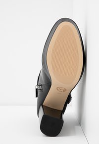 Scarpa alta nera in pelle con una finitura liscia, punta quadrata e suola in gomma testurizzata con scanalature orizzontali. Due dettagli decorativi.