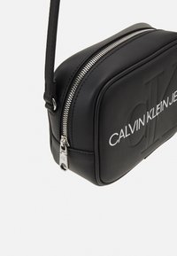 Černá obdélníková crossbody taška Calvin Klein Jeans se stříbrným zipem a embosovaným logem na předním panelu.