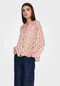 Blouse à manches longues rose clair avec motif paisley, col à volants et fermeture par boutons, associée à un jean large bleu foncé.