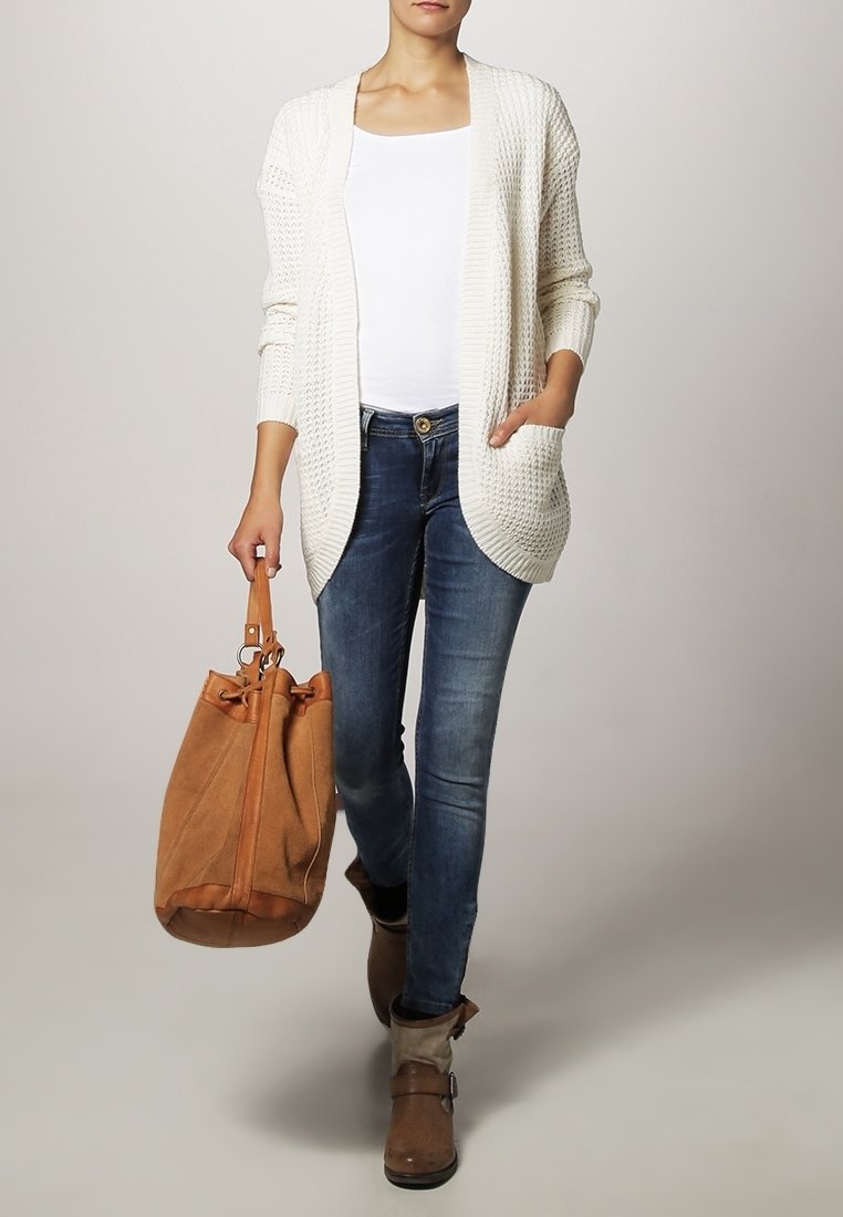 Femme portant un cardigan en maille blanc, un haut blanc, un jean bleu, des bottines marron, tenant un grand sac à main en daim marron dans la main droite.