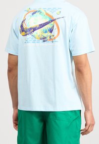 Světle modré tričko vyrobené z bavlny s barevným motivem na zádní straně, který zobrazuje globus, palmy a velký Nike swoosh.