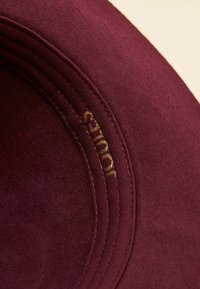 Pălărie din fetru burgundy cu broderie a logo-ului în culoarea aurie. Prezintă o textură netedă și un detaliu cu bandă stratificată, evidențiind măiestria meticuloasă.
