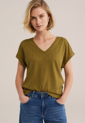Mujer rubia con pendientes de aro dorados, camiseta de manga corta con escote en V texturizada de color verde oliva y vaqueros azules con las manos en los bolsillos, fondo neutro.