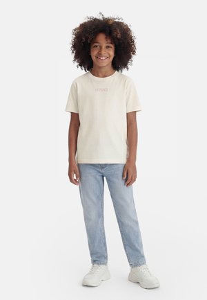 Παιδί με σγουρά μαλλιά που χαμογελά, φοράει κρεμ T-shirt Versace, ανοιχτό μπλε τζιν και λευκά αθλητικά παπούτσια, στέκεται μπροστά σε ένα λευκό φόντο.