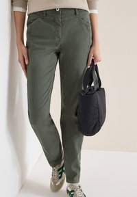 Personne portant un pantalon vert olive et des baskets beiges à rayures vertes, tenant un petit sac fourre-tout noir par ses poignées.