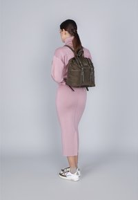 Mochila de nailon verde olivo oscuro con múltiples bolsillos con cremallera, correas ajustables y un diseño estructurado, llevada sobre un vestido rosa.