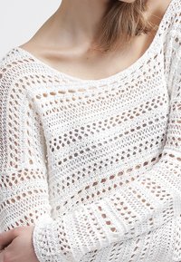 Pull en tricot blanc avec un design ajouré et des rayures horizontales. La texture est aérienne, avec des manches longues et un décolleté large.