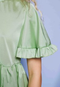 Svetlo zelena satenasta obleka z napihnjenimi kratkimi rokavi in naborki na manšetah, ki jo nosi oseba s svetlo kožo na svetlo modri ozadju.