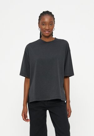 Νεαρή γυναίκα με πλεξούδες, ντυμένη με μαύρο χαλαρό μπλουζάκι με κοντά μανίκια και μαύρο παντελόνι, στέκεται μπροστά σε απλό λευκό φόντο.