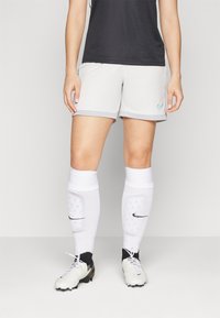 Graue Sportshorts mit einem türkisfarbenen Logo, kombiniert mit weißen Socken und Schuhen mit schwarzen Akzenten. Glattes Material und enganliegendes Design.