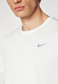 White short-sleeve Nike tee with a textured pattern, featuring a grey logo on the left chest area and a classic round neckline.