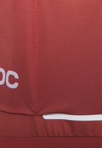 Czerwony materiał sportowy o gładkiej teksturze z poziomym białym paskiem na dole i kontrastującymi pomarańczowymi szwem.
