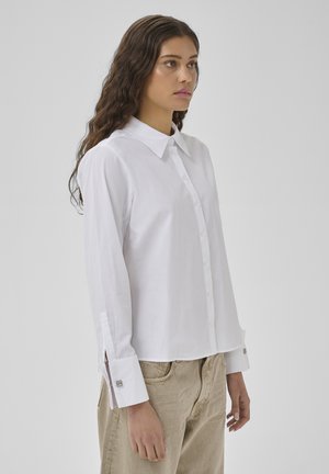 Kvinde med langt bølget hår iført en hvid skjorte med knapper og beige bukser, står og kigger til siden mod en ensfarvet baggrund.