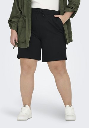 Kvinne iført svarte shorts med snor i midjen, hvite joggesko og en olivengrønn jakke, stående med én hånd i lommen.