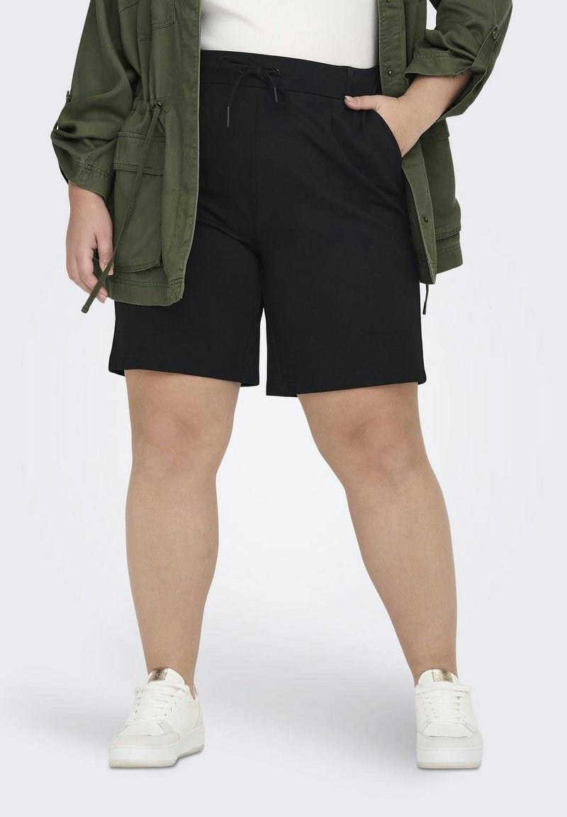 Žena oblečená v černých šortkách na stažení, bílých teniskách a olivově zelené bundě, stojící s jednou rukou v kapse.