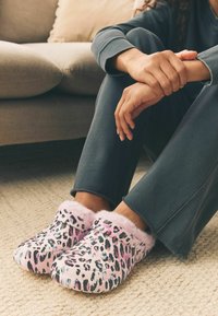 Papuci roz cu imprimeu de leopard, cu căptușeală moale și pufoasă. Formă rotundă, concepuți pentru confort. Purtați cu pantaloni negri, cu croială lejeră, pe o suprafață texturată.