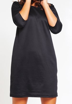 Vrouw draagt een zwarte jurk tot op de knieën met driekwart mouwen, staand tegen een effen witte achtergrond, handen dicht bij haar gezicht.