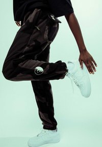 Joggers de forro polar en camuflaje negro y gris con puños elásticos, bolsillos laterales y un logo redondo. Combinados con zapatillas Nike Air Force 1 blancas.