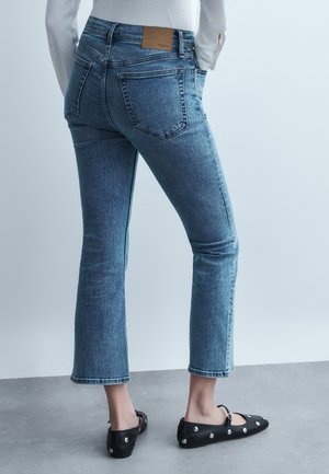 Vue arrière d'une personne portant un jean bleu court avec des poches arrière et des ballerines noires ornées de clous argentés sur un sol en béton.