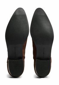 Un paio di scarpe da cerimonia in suede marrone viste dal basso, con suole in gomma nera caratterizzate da sottili scanalature orizzontali e il marchio "Tommy Hilfiger".