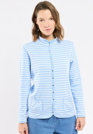 Femme portant un cardigan à rayures bleu clair et blanches avec des poches avant et un pantalon bleu, sur un fond blanc uni.