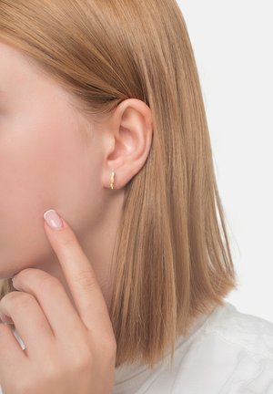 Närbild av en kvinnas vänstra öra med kort blont hår, som bär en liten gyllene snurrad hoopörhänge, medan hon rör vid sin kind med ett manikyrerat finger.